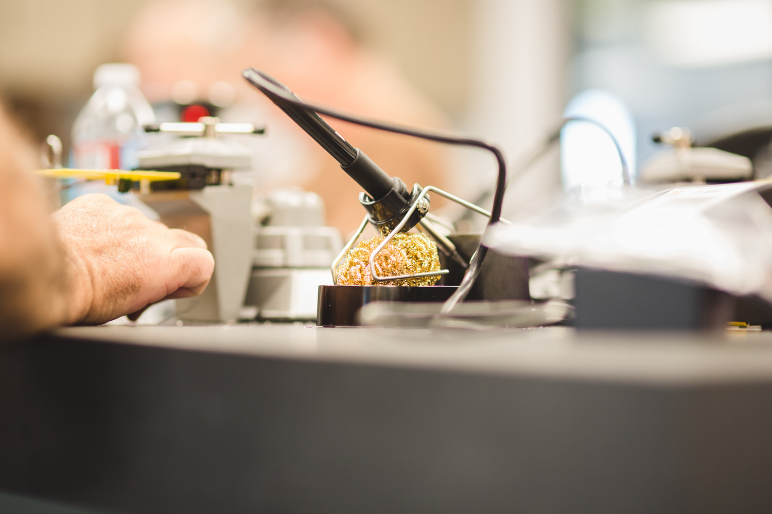 A close up of a student’s hand resting on the table near a soldering iron set in its station. A soldering iron is a handheld tool with an insulated handle and heated metal tip used to melt solder. 