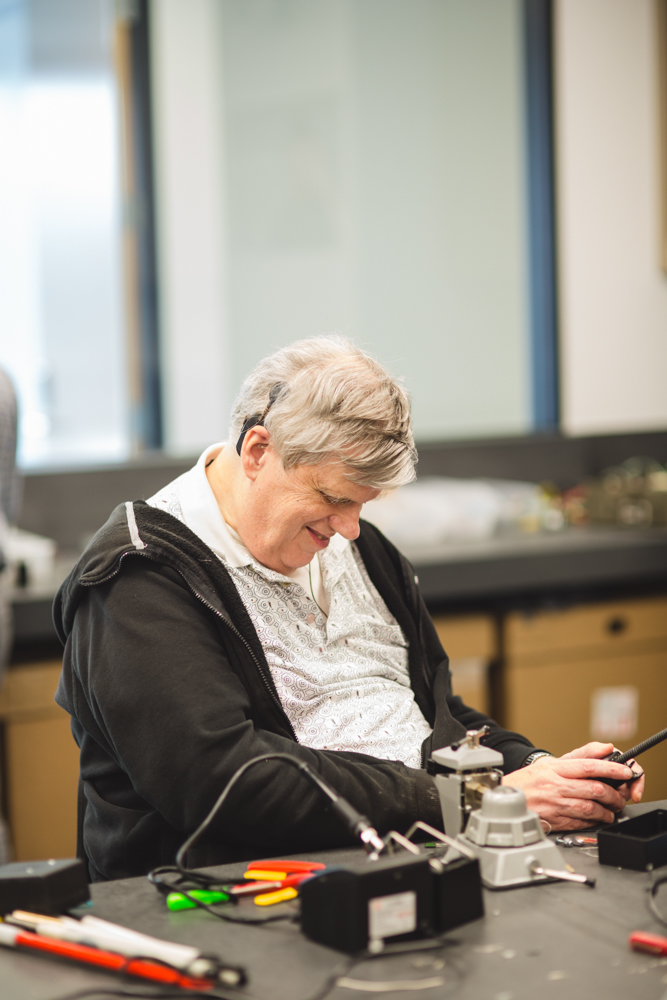 A smiling grey-haired male student wearing a black hoodie and a white button-up sits at the table grasping a completed continuity tester.