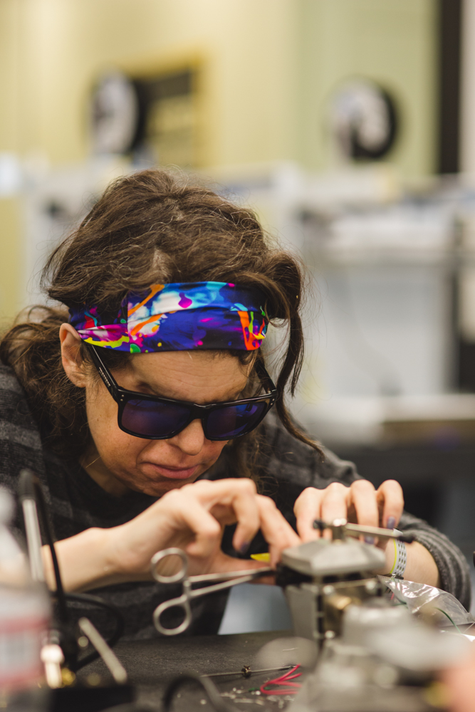 A female soldering student wearing reflective sunglasses and a colorful headband leans over her work station, deep in a concentration. A steel vice is used to steady a yellow circuit board for ease of work while soldering. 