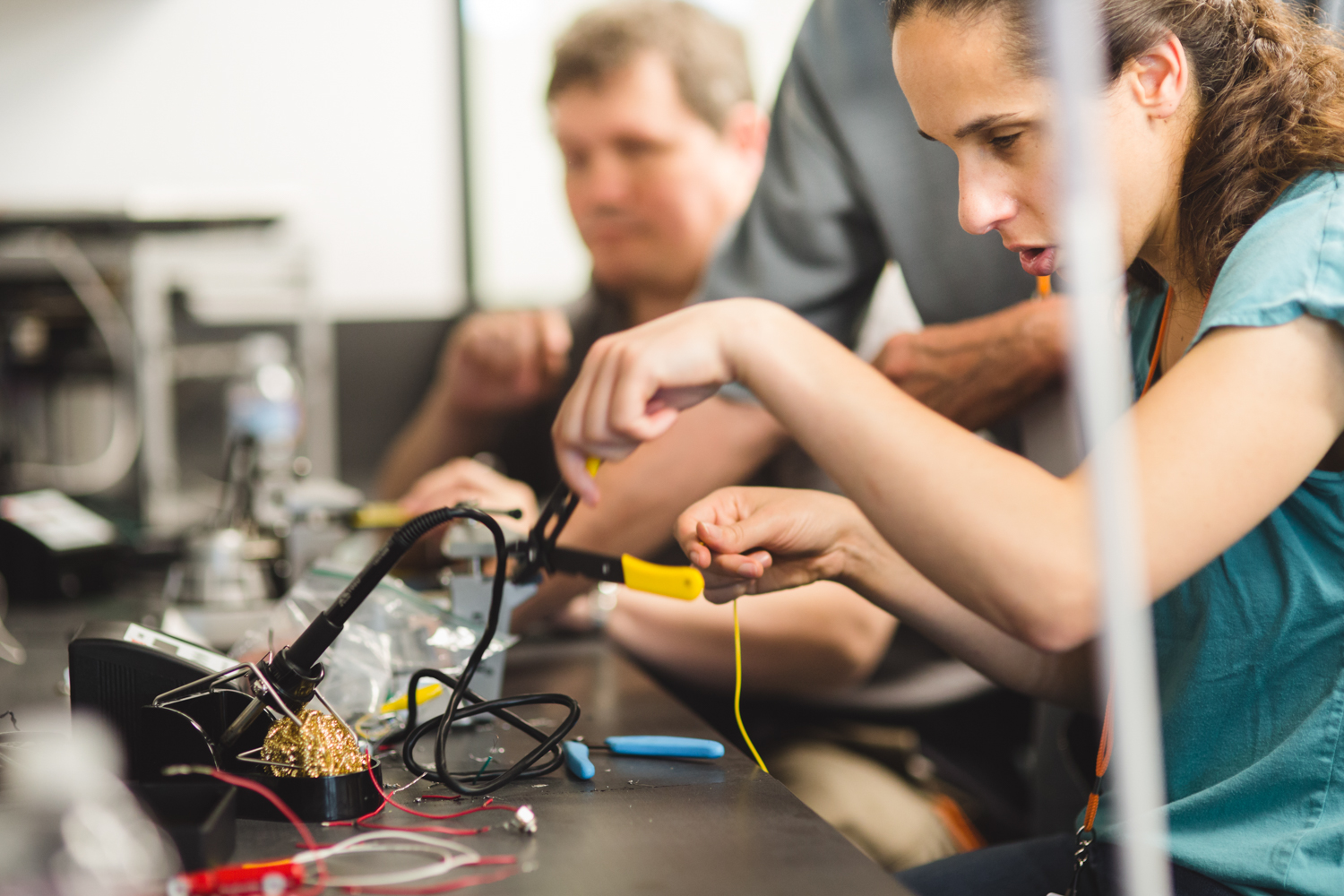 Clasping a pair of yellow wire-strippers, a female student in a teal shirt uses the instrument’s notched jaws to remove the insulation from a section of yellow wire. Her other tools are scattered on the table in front of her. Other students are hard at work in the background. 