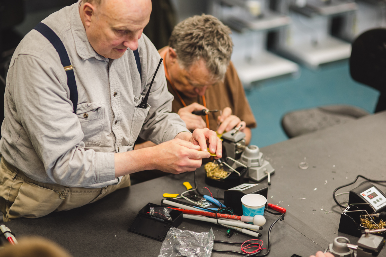 A man in a grey shirt and suspenders clasps a yellow circuit board. Behind him, the student with the tawny shirt is hard at working with his soldering iron in hand. A smattering of tools are sprawled across the table next to a folded cane. 