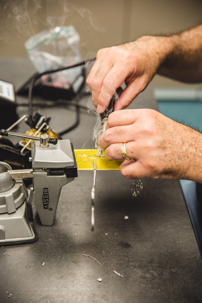 A curl of smoke rises from the tip of a hot soldering iron as a student melts points of solder onto his circuit board. 