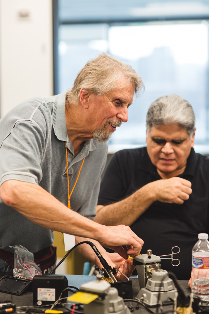 Two older male students collaborate at a busy soldering station. 