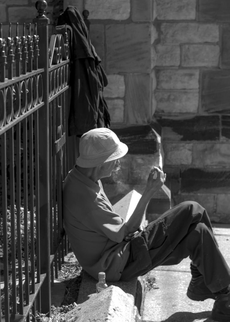 Photograph 1: A man sits on a concrete ledge and leans his weight into wrought iron fence. His wears a bucket hat and the smoke from the cigarette curled in his right hand catches in the light. A bottle of hard liquor is perched next to him on the ground, slightly concealed by an angular concrete block. Click the image to hear the corresponding audio file.