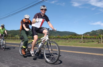 Two men pedal a tandem bike whilst giving the camera a thumbs up.