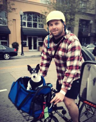 Sam pedals by dressed in a flannel. He is accompanied by a dog in his bike's basket.