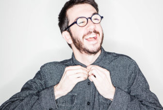 Communications director Will Butler laughs as he buttons the top of his shirt in front of a white background