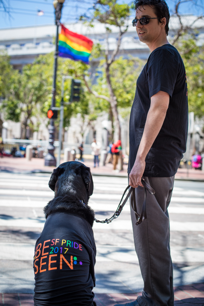 Ethan and his guide dog Gershwin show off their LightHouse Pride t-shirts across from the UN Plaza, with a rainbow flag billowing in the background.