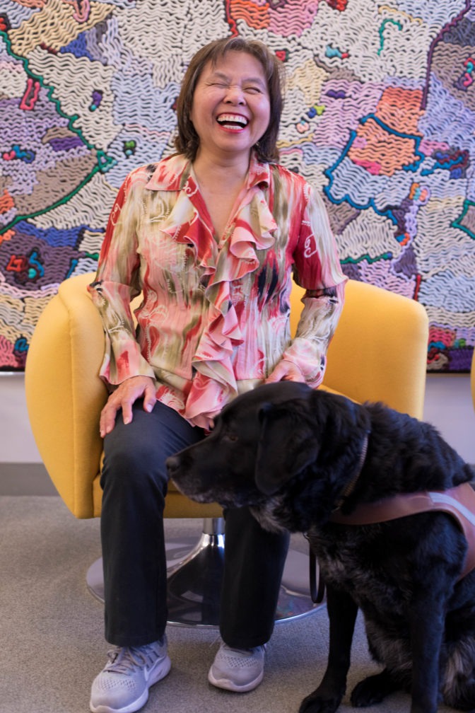 Kit Lau smiles with her guide dog Alisa in front of a colorful background.