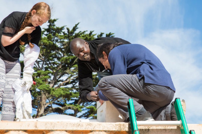 Ojok and Lowell student Ellie show Ahmet the ropes as he feels a beehive box.