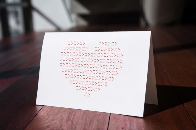 Red ink-print and braille X's and O's form the shape of one large heart on a white greeting card. The inside of the card is blank.