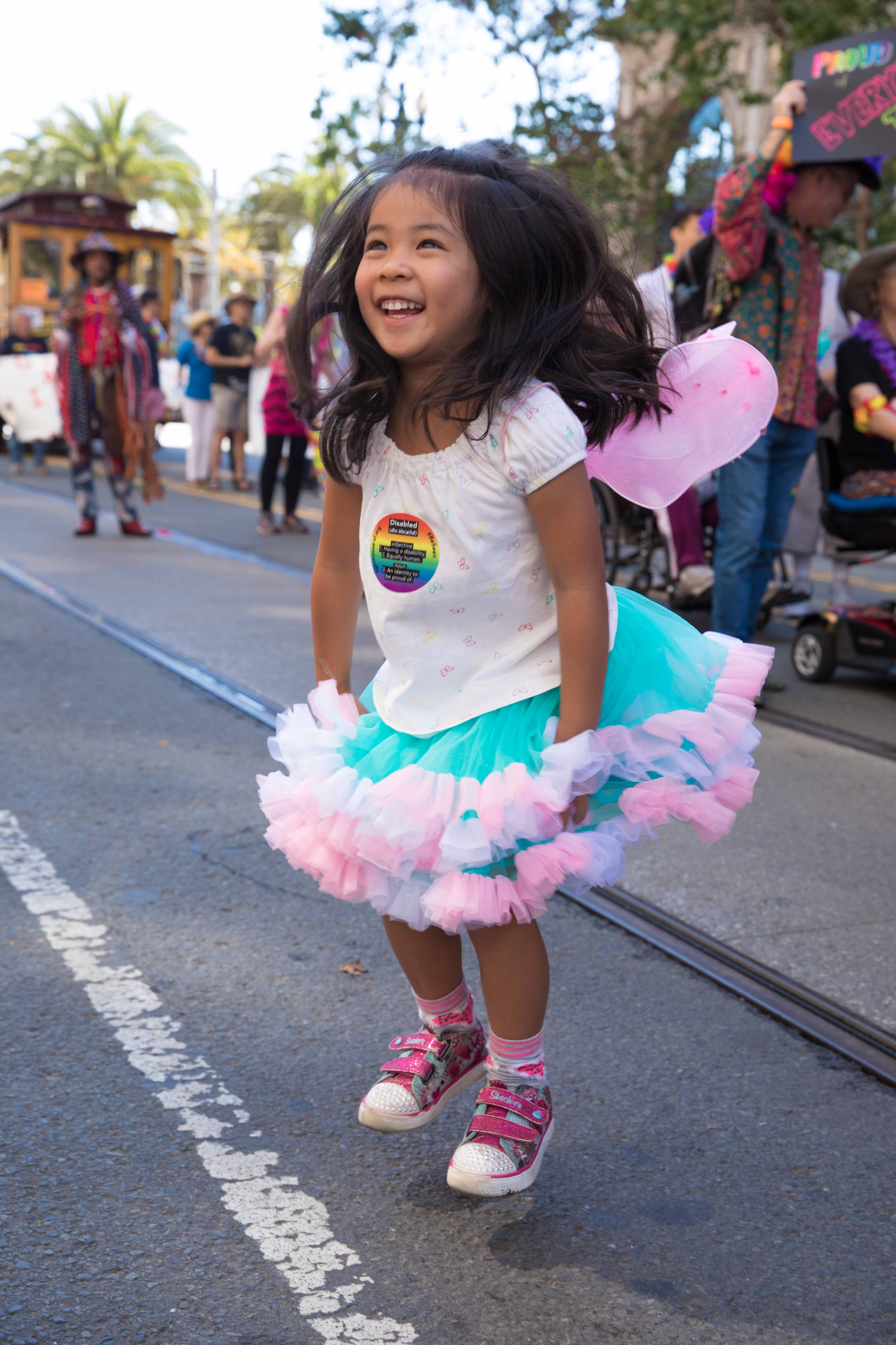 A little girl wearing a tutu and fairy wings smiles and jumps into the air.