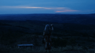A woman stands on a ledge in front of a fresh night sky with low, blue light in front of an open frontier. She holds her phone up to photograph the scene.