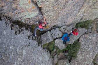 A climber ascends a free-standing rock, while two people stand on a rock below her.