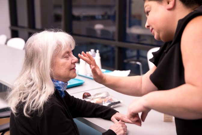 At left, an older woman sits with her eyes closed. At right, a makeup approaches the tip of the woman's nose with a delicate hand to apply concealer.