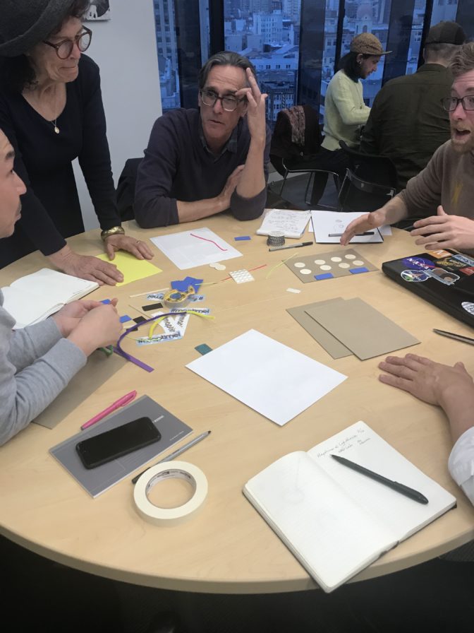 Five Maptimers sit around a circular table while creating their tactile maps. Their tools are pens, paper, foam core, glue, scissors, wiki sticks — anything you can add to paper to build out a tactile layout.