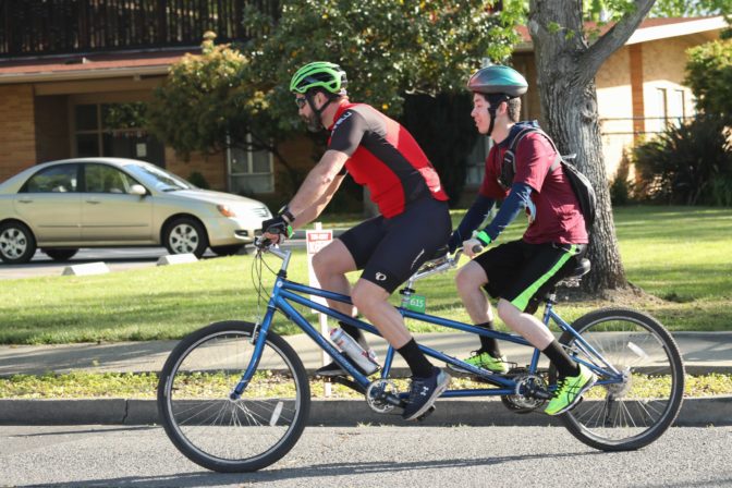 Volunteer Matt, a middle aged man, and Danny, who is teenaged and blind, ride together on a tandem bicycle.