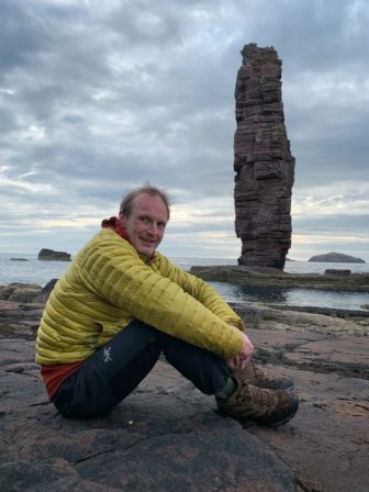 Red sitting on a rocky beach at Sandwood Bay, on the far north-west coast of mainland Scotland, with Am Buachaille towering behind him.