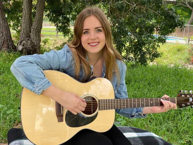 Meghan Downing sits outside on a black and white blanket and smiles while she strums a guitar.