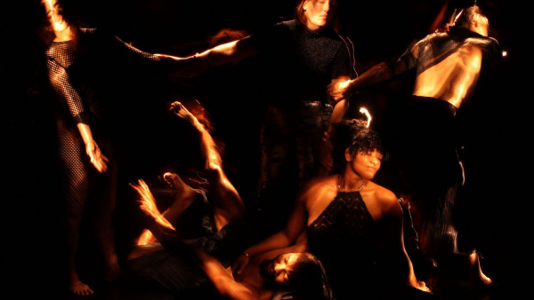 A photo from Into The Dark. Against a dark background, five figures appear, their bodies lit as if by a handheld flashlight. Three in the background link arms leaning away from each other as if in despair, or maybe rapture, while one figure sits on the ground in front, cradling the head of the fifth, prone figure, in her lap as the arm of the figure outstretched on the floor reaches up toward the trio standing behind