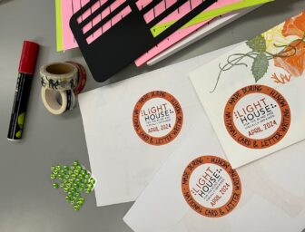 Art and adaptive letter writing supplies on a table with sealed envelopes and “Made During National Card & Letter Writing Month Lighthouse April 2024” round stickers on the back.