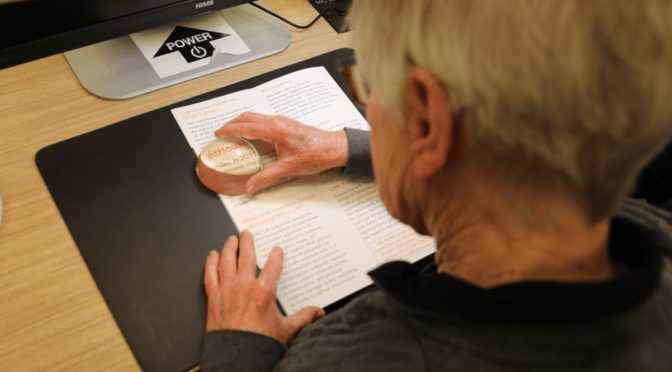 Seen from behind, a student sits at a desk and uses a dome magnifying glass to read text