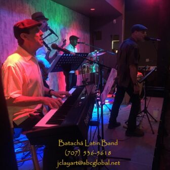 The Batacha Latin Band in performance consisting of a keyboardist, guitarist, drummer and lead vocalist onstage singing into microphones as they play their instruments. On the photo is contact info: 707-536-5618 and jclayart@sbcglobal.net.
