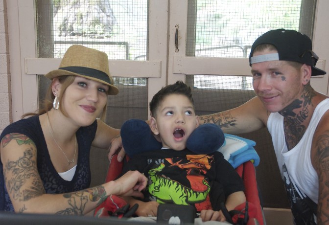 young boy poses, mouth agape, parents stylish between father's tattoos and mother's fedora