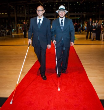 two men in navy suits with canes walk down the red carpet, one wearing a fedora
